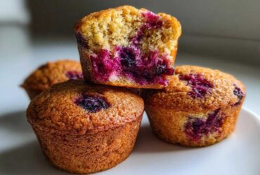 A stack of golden brown blueberry protein muffins, with the top one cut in half showing the moist interior.