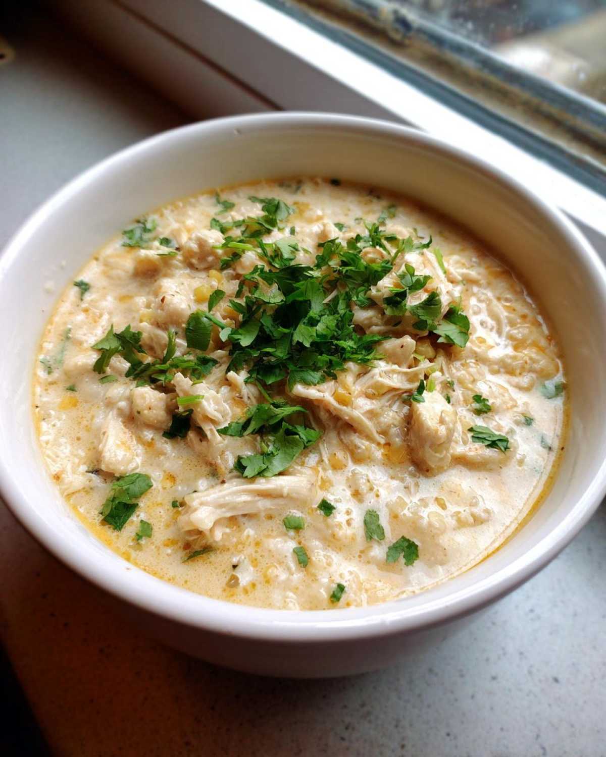 A close-up of a white bowl filled with creamy, shredded cream cheese chicken chili, garnished generously with fresh cilantro.
