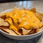 A white bowl filled with tortilla chips generously topped with bright orange nacho cheese sauce.