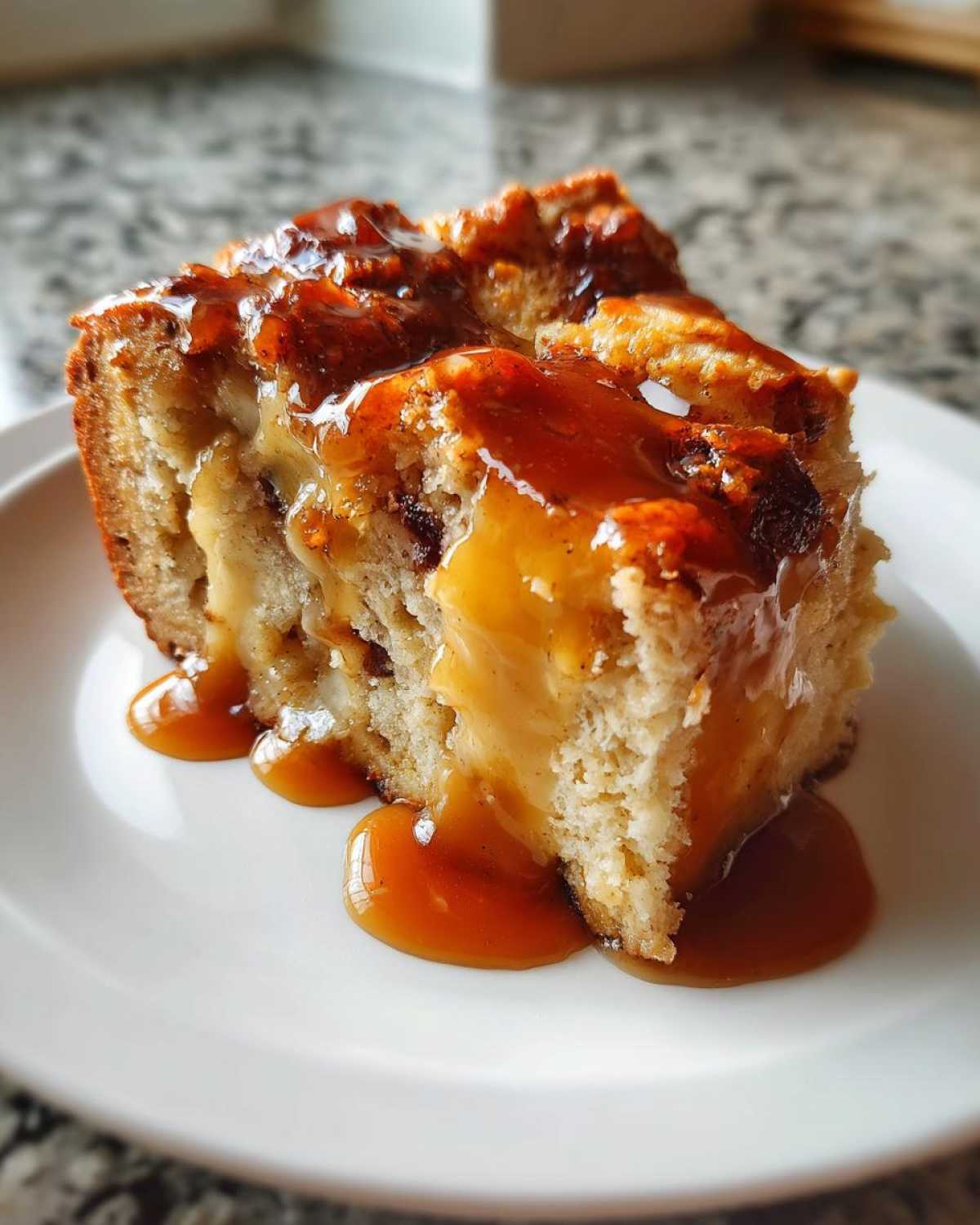 A close-up of a rich slice of bread pudding with bourbon sauce generously drizzled over the top and pooling on the white plate.