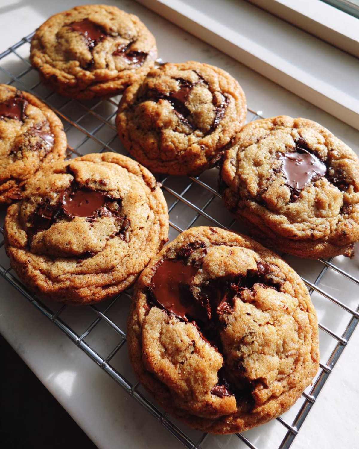 Six golden brown butter chocolate chip cookies cooling on a wire rack with melted chocolate centers.