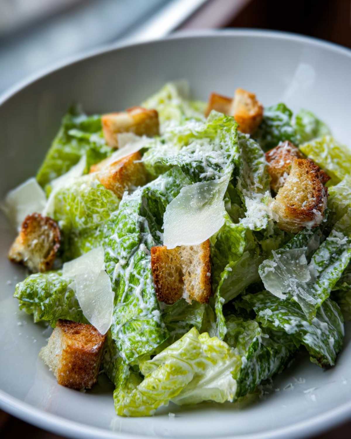 A close-up shot of a fresh caesar salad in a white bowl, featuring crisp romaine, creamy dressing, golden croutons, and shaved Parmesan cheese.