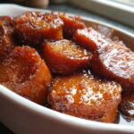 Close-up of thick-cut candied sweet potatoes glistening in a rich, dark glaze served in a white oval dish.