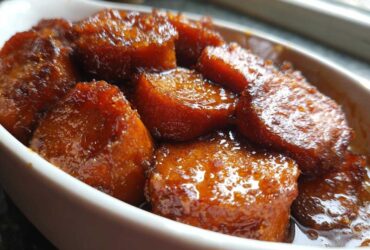 Close-up of thick-cut candied sweet potatoes glistening in a rich, dark glaze served in a white oval dish.