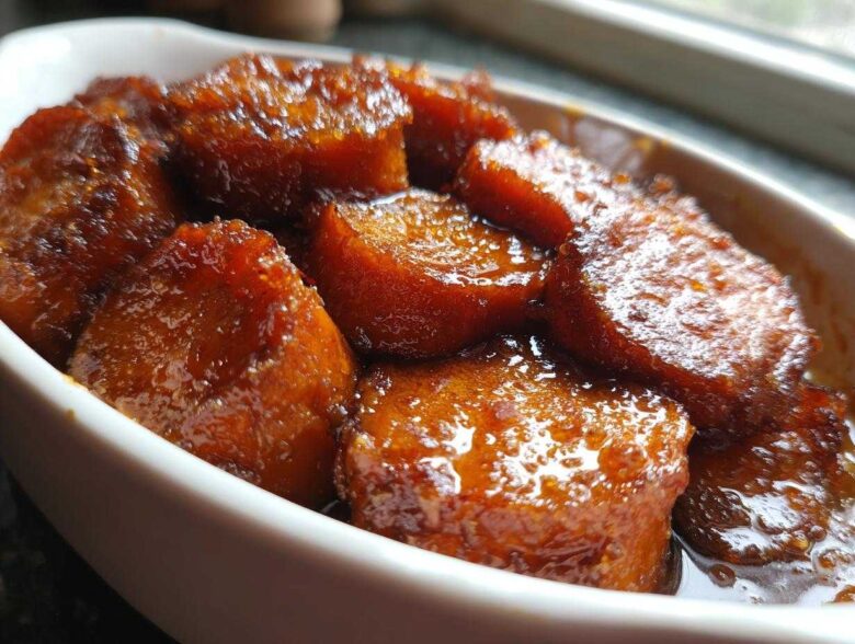 Close-up of thick-cut candied sweet potatoes glistening in a rich, dark glaze served in a white oval dish.