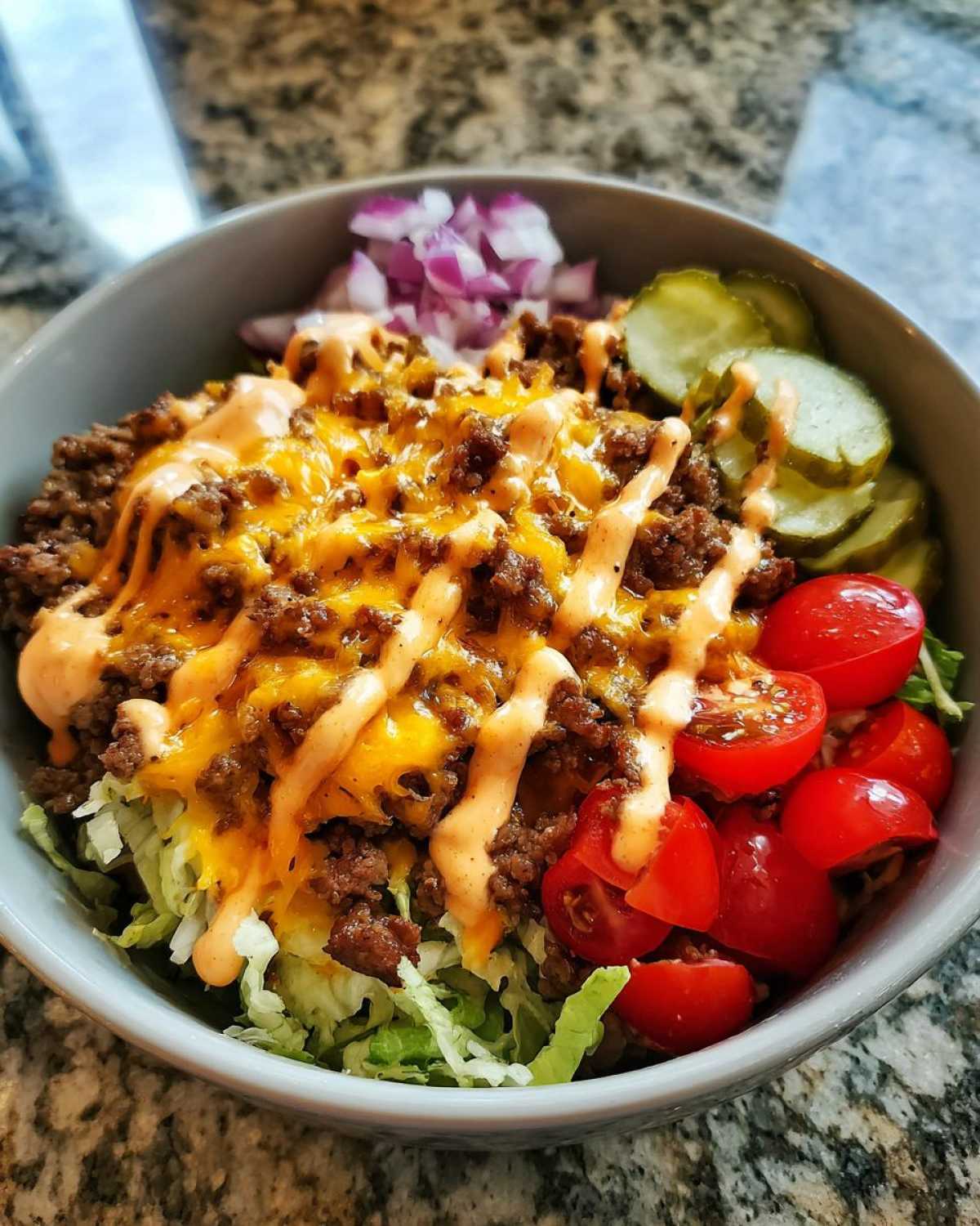 A close-up view of a fully loaded cheeseburger bowl featuring seasoned ground beef, melted cheese, special sauce, lettuce, pickles, tomatoes, and onions.