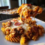 A fork lifts a cheesy, saucy portion of pizza casserole, showing long cheese pulls over rotini pasta and pepperoni.