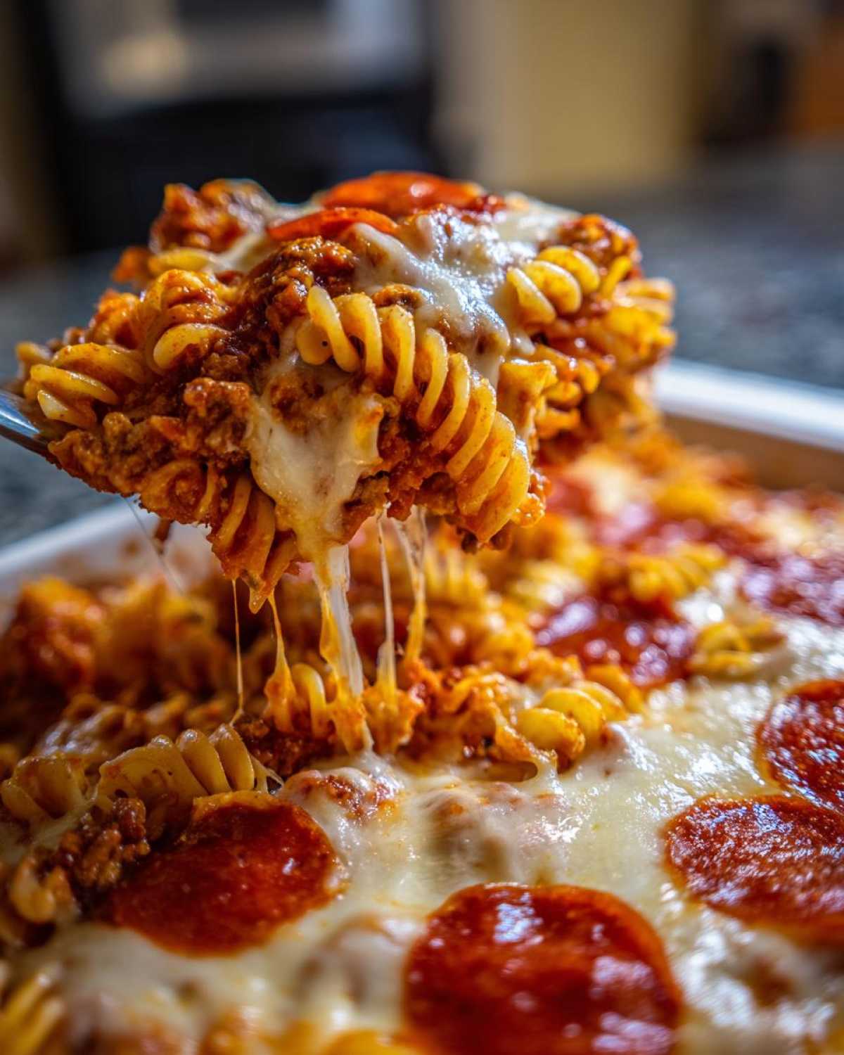 A serving spoon lifts a portion of cheesy pizza casserole made with rotini pasta and pepperoni.