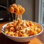 A spoonful of creamy, cheesy enchilada pasta being lifted from a white bowl, showing long cheese pulls.
