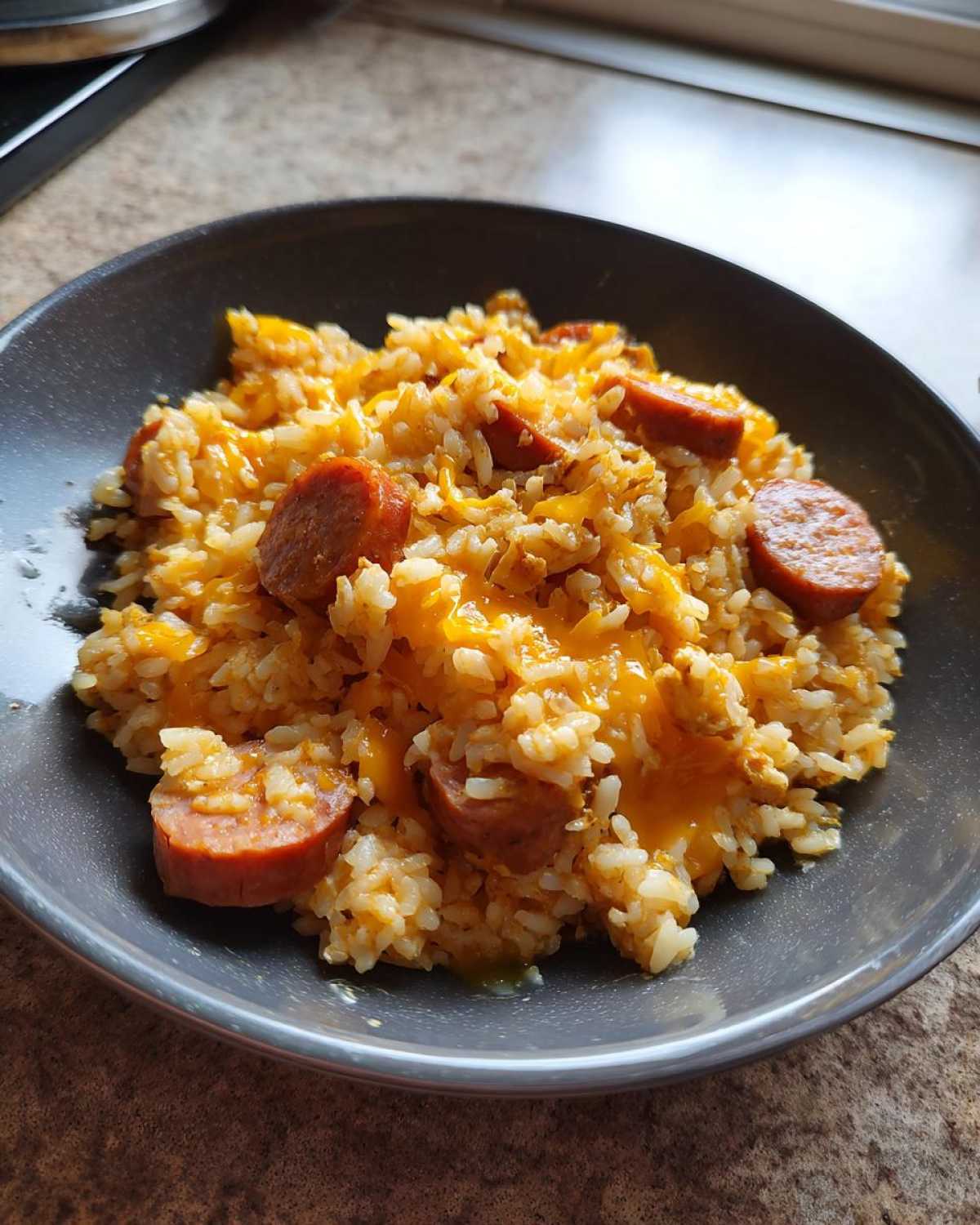 Close-up of cheesy sausage and rice mixed together and served in a dark grey bowl.