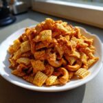 A white bowl piled high with savory, seasoned chex party mix pieces and whole roasted cashews.
