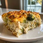 A square serving of creamy chicken broccoli rice casserole topped with golden, melted, and slightly browned cheese.