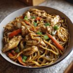 Close-up of a bowl filled with savory chicken chow mein recipe, featuring noodles, sliced chicken, carrots, and green onions.