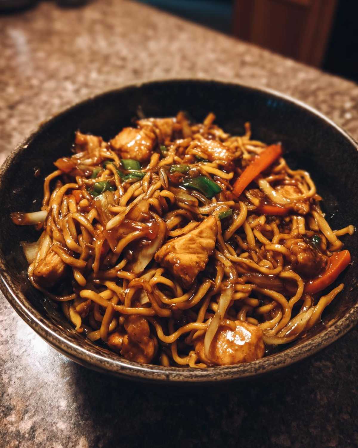 Close-up of a dark bowl filled with savory chicken chow mein recipe, featuring noodles, chicken pieces, and vegetables.