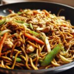 Close-up of delicious chicken chow mein recipe stir-fried with noodles, chicken pieces, carrots, and green onions in a black wok.