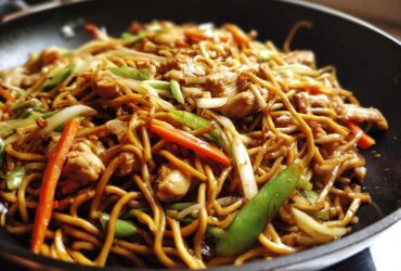 Close-up of delicious chicken chow mein recipe stir-fried with noodles, chicken pieces, carrots, and green onions in a black wok.