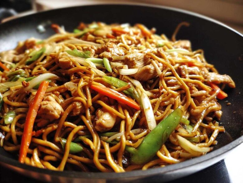 Close-up of delicious chicken chow mein recipe stir-fried with noodles, chicken pieces, carrots, and green onions in a black wok.