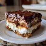 A close-up of a layered slice of chocolate eclair cake with graham cracker layers, creamy filling, and thick chocolate frosting.