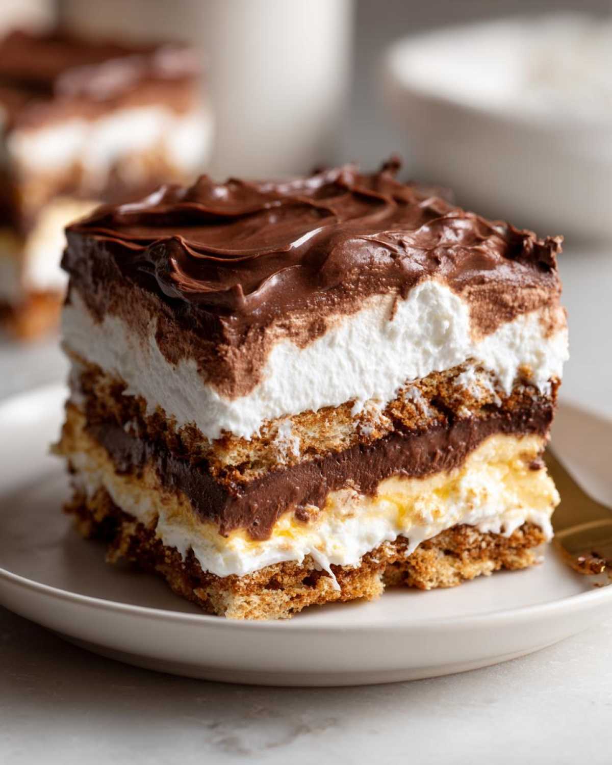 A perfect square slice of chocolate eclair cake showing layers of graham crackers, pudding, cream, and chocolate frosting.