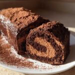 A rich, chocolate yule log cake dusted heavily with cocoa powder, with one slice cut to show the swirl of filling.