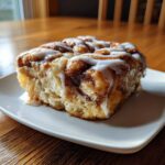 A close-up of a warm slice of cinnamon roll casserole topped generously with white icing.