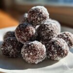 A pyramid stack of dark chocolate bourbon balls rolled in white coconut flakes, sitting on a white plate.