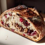 A close-up, sunlit cross-section of homemade cranberry walnut bread showing a beautiful open crumb studded with bright red cranberries and walnuts.