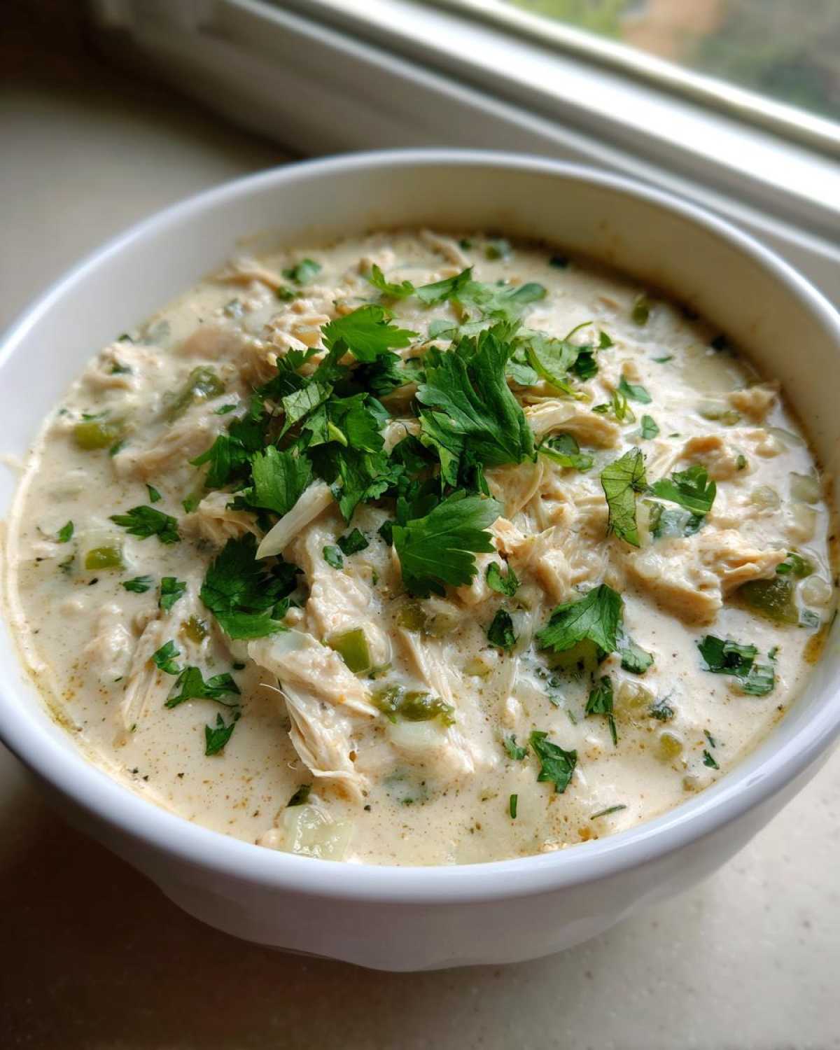 A close-up of a white bowl filled with rich, creamy cream cheese chicken chili, topped with fresh green parsley.