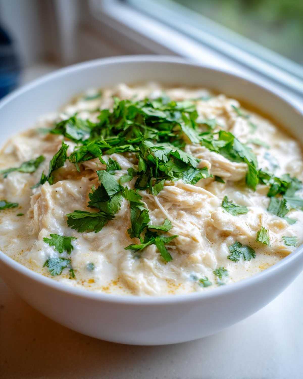 A close-up of a white bowl filled with rich, creamy white cream cheese chicken chili, topped generously with fresh cilantro.
