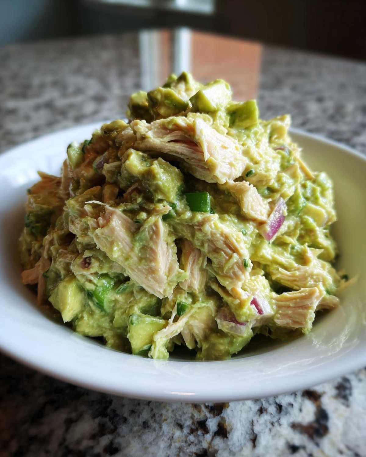 A close-up of creamy avocado chicken salad, featuring shredded chicken and chunks of avocado, served in a white bowl.