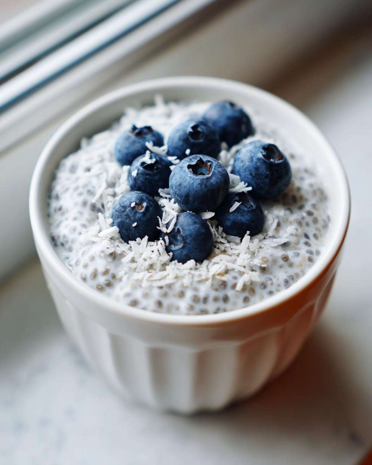 A white bowl of creamy coconut chia pudding topped with fresh blueberries and shredded coconut.