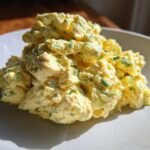 A close-up of a generous mound of creamy, yellow egg salad recipe speckled with green chives on a white plate.