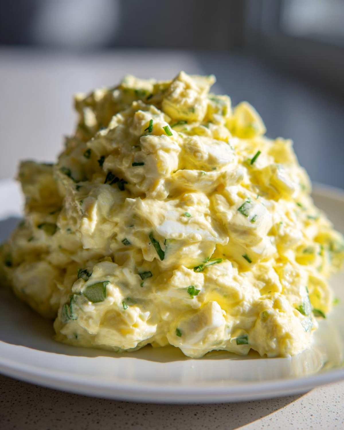 A close-up, brightly lit mound of creamy egg salad recipe mixed with visible chunks of egg and sprinkled with fresh green chives.