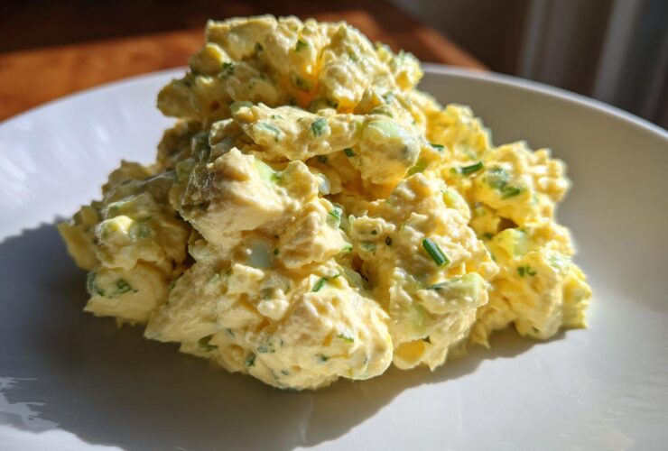 A close-up of a generous mound of creamy, yellow egg salad recipe speckled with green chives on a white plate.