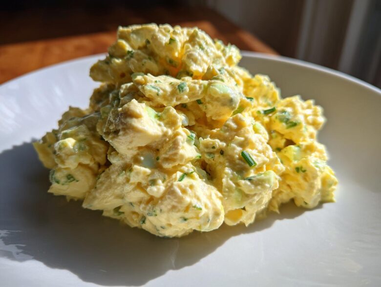 A close-up of a generous mound of creamy, yellow egg salad recipe speckled with green chives on a white plate.
