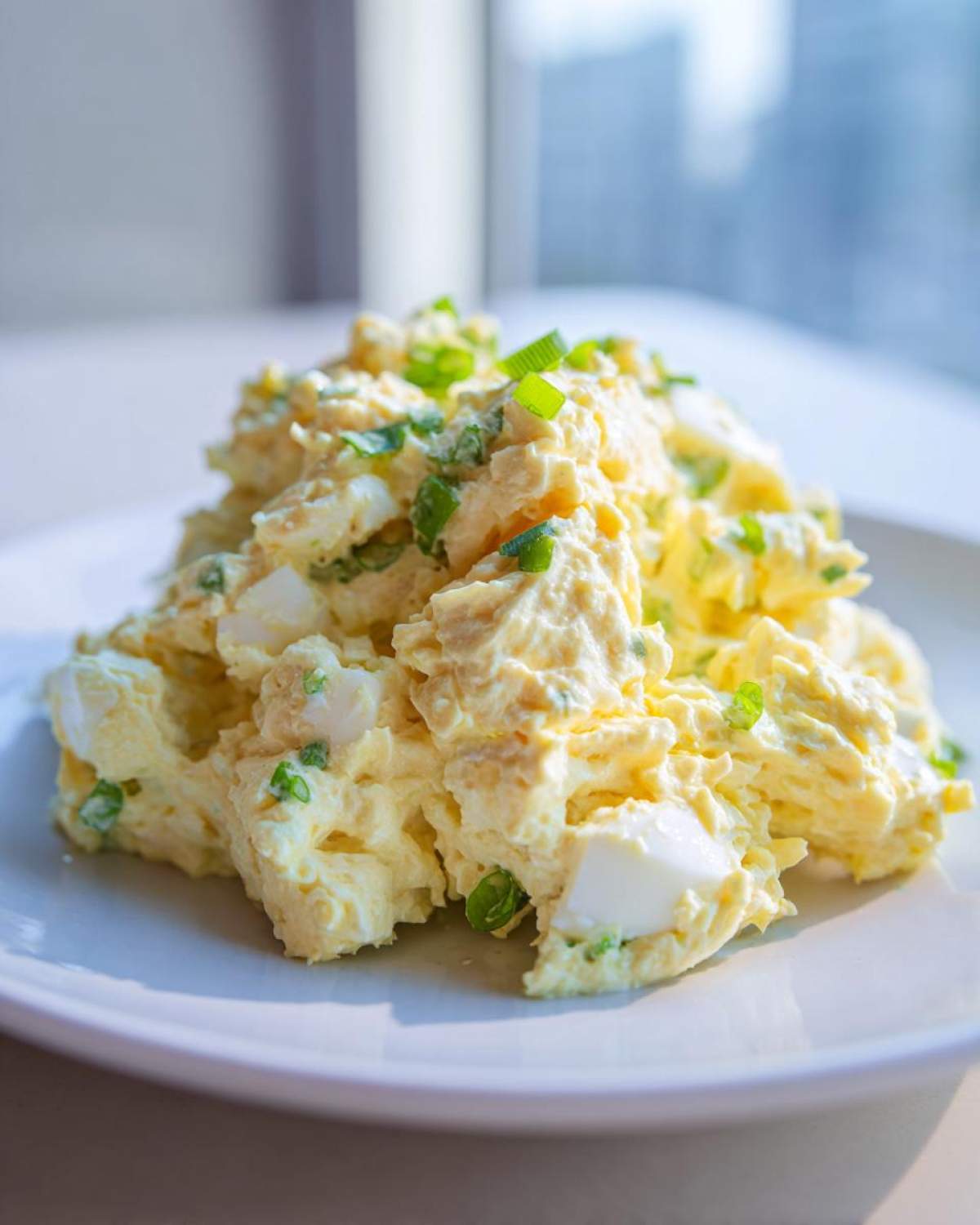 A generous serving of creamy egg salad recipe topped with chopped green onions, sitting on a white plate.