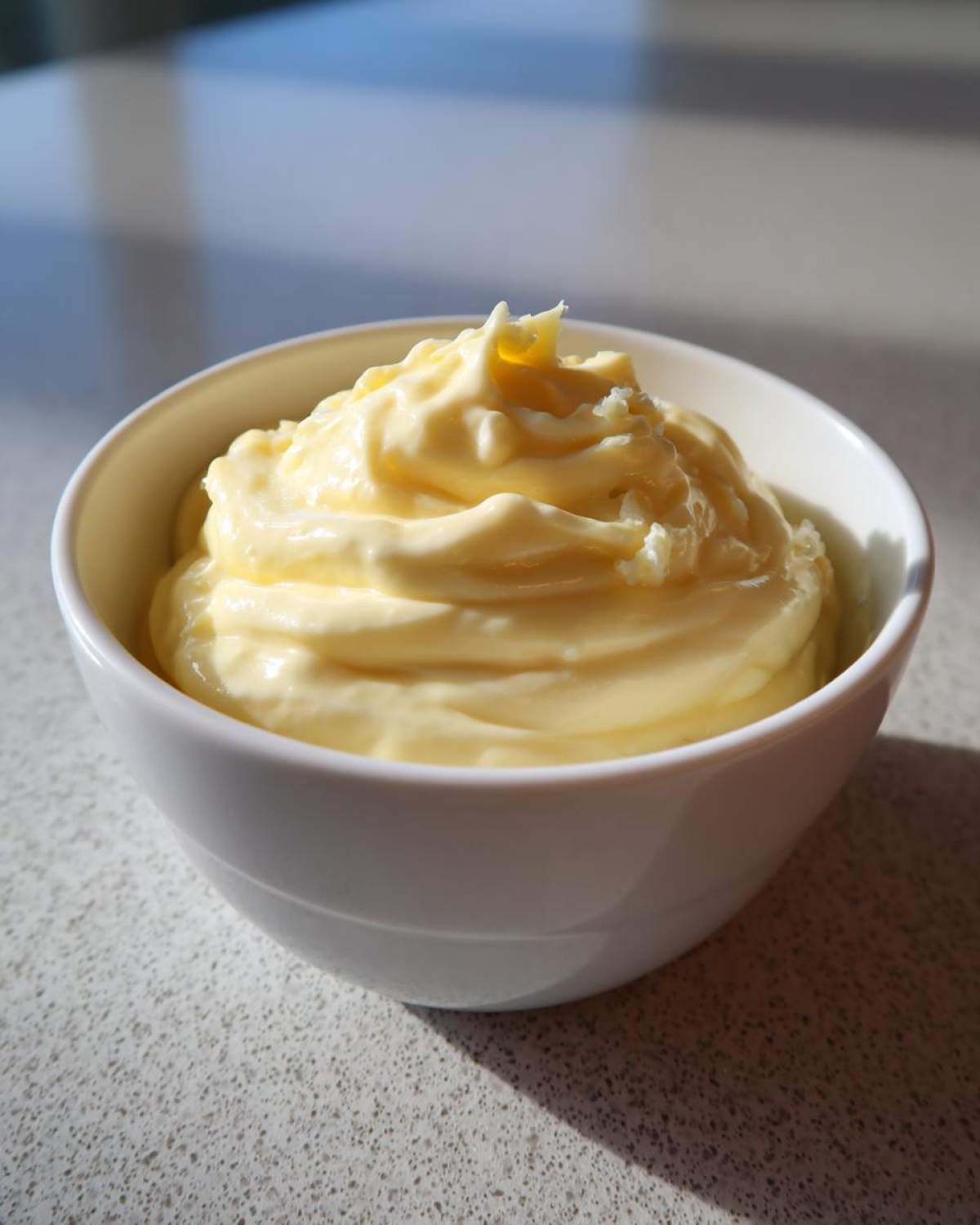 A close-up of rich, pale yellow, creamy garlic aioli swirled high in a small white bowl.