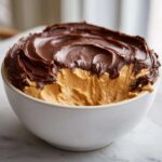 Close-up of a white bowl filled with creamy peanut butter dip, swirled with a thick layer of chocolate topping.