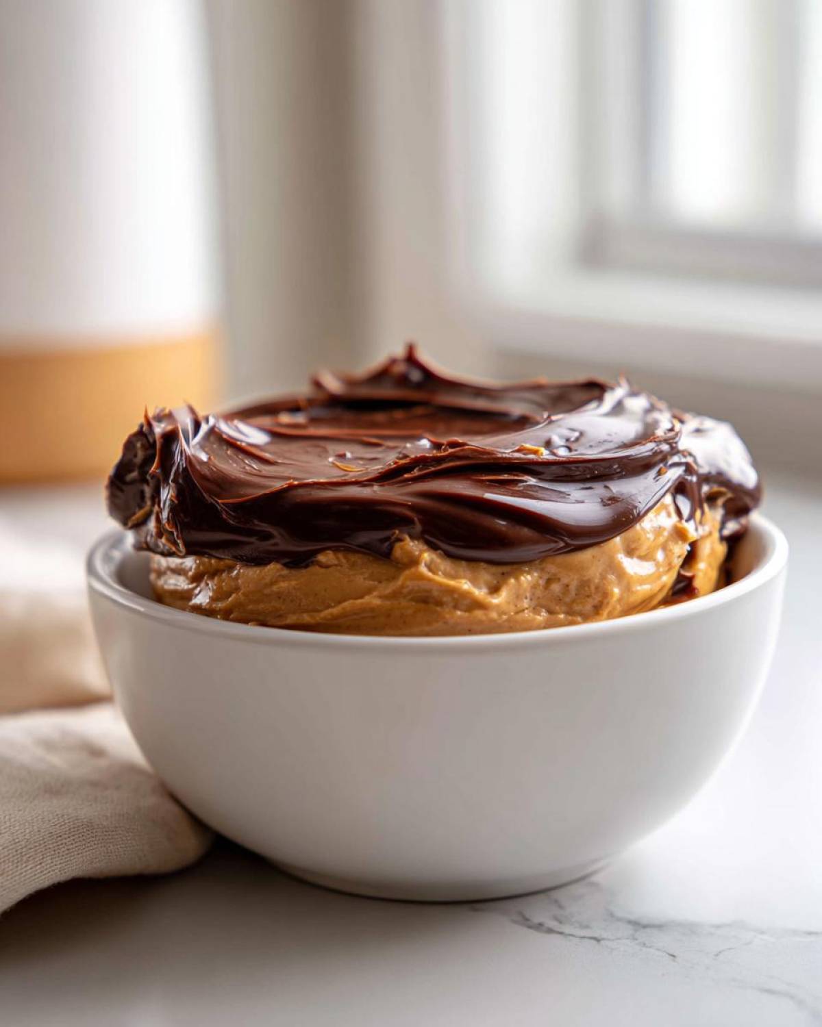 Close-up of a white bowl filled with creamy peanut butter dip topped with melted chocolate.