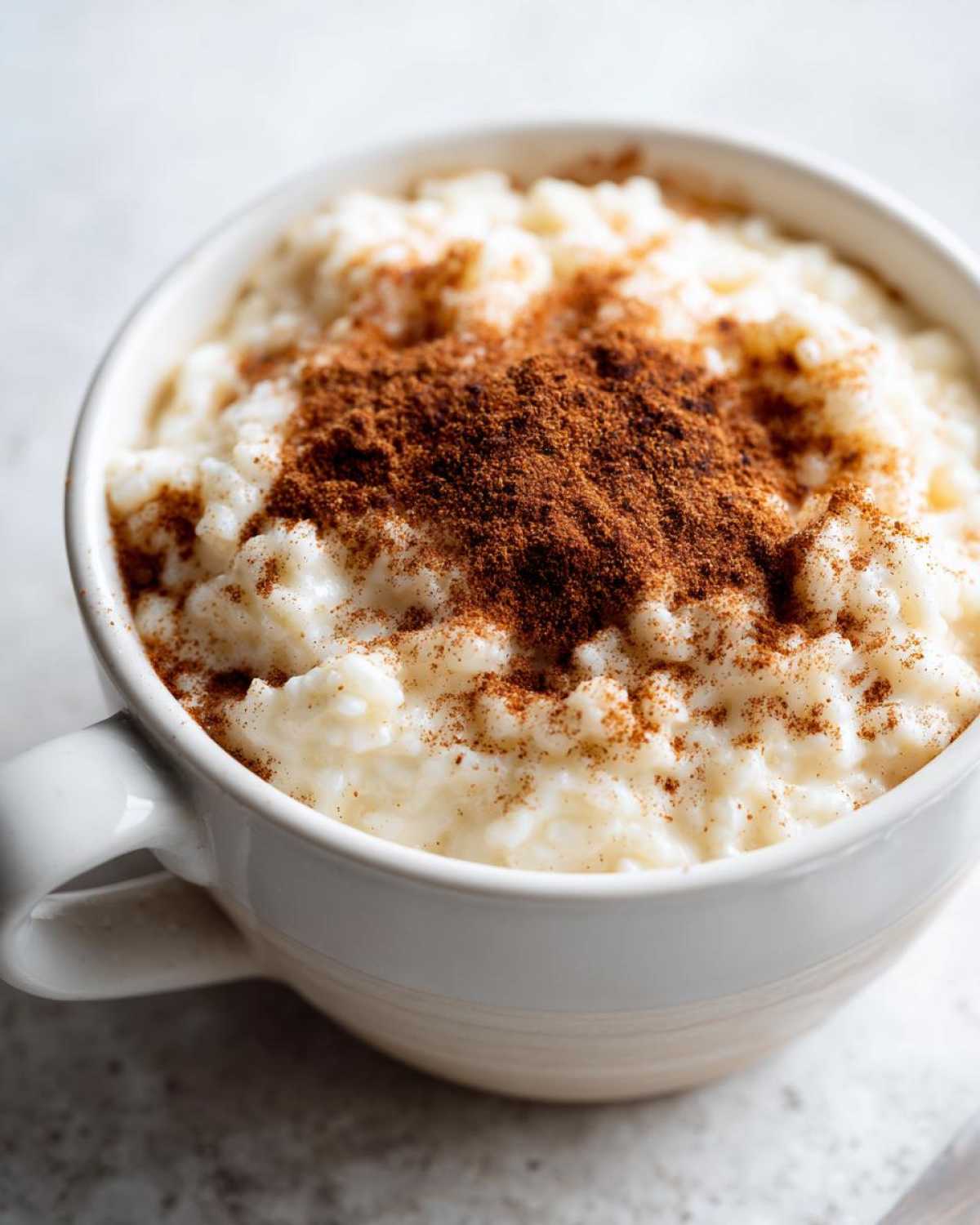 Close-up of creamy rice pudding served in a white mug, generously dusted with ground cinnamon.