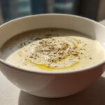 A close-up of a bowl of creamy roasted cauliflower soup, drizzled with olive oil and sprinkled with pepper and herbs.