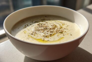 A close-up of a bowl of creamy roasted cauliflower soup, drizzled with olive oil and sprinkled with pepper and herbs.
