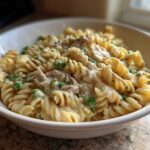A close-up of creamy tuna pasta made with fusilli noodles and green peas served in a white bowl.