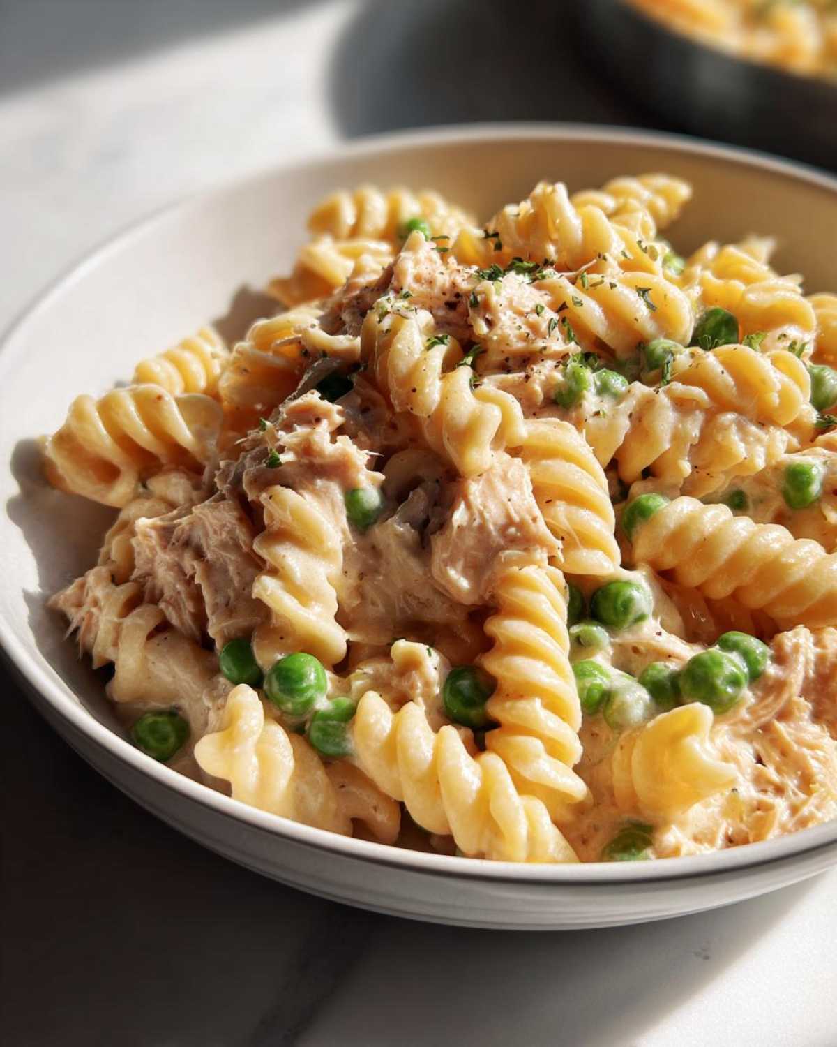 Close-up of creamy tuna pasta made with rotini noodles and bright green peas, garnished with herbs.