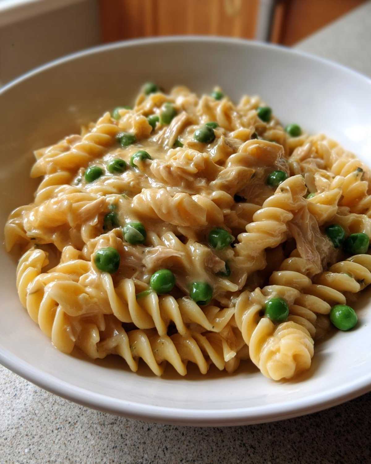 A close-up of creamy tuna pasta made with rotini noodles and bright green peas served in a white bowl.