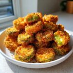 A mound of golden brown, crispy Fried Pickles piled high on a white plate, one showing the bright green interior.