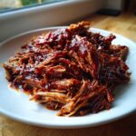 Close-up of tender, shredded crock pot pulled pork coated in rich barbecue sauce on a white plate.