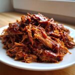 A generous serving of saucy, shredded crock pot pulled pork piled high on a white plate.