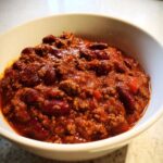 Close-up of a white bowl filled with rich, thick crockpot chili featuring ground meat and dark red kidney beans.