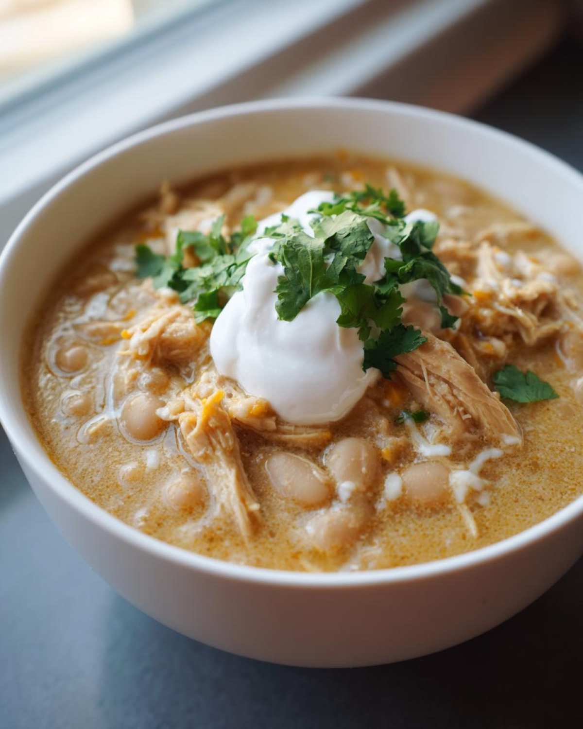 A close-up of a white bowl filled with creamy crockpot white chicken chili, topped with sour cream and fresh cilantro.
